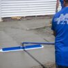 Worker in a blue shirt smoothing a freshly poured concrete driveway with a long blue-handled trowel attachment.