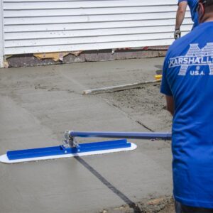 Worker in a blue shirt smoothing a freshly poured concrete driveway with a long blue-handled trowel attachment.