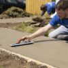 Worker smoothing wet concrete with a trowel on a new sidewalk; another worker is in the background.
