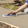 Person smoothing freshly poured concrete with a blue-handled float trowel on a construction site surface