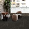 Two woven wicker chairs with dark cushions face a round wicker table on a stone-floored balcony with a potted plant nearby and urban buildings in the background,”