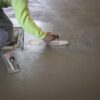 Construction worker in a neon-green sleeve smooths wet concrete with a hand trowel and edging tool on a floor slab