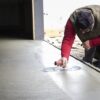 Worker smoothing a freshly poured concrete surface with a hand trowel, wearing a red long-sleeve shirt and olive vest outdoors at a construction site