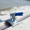 Close-up of a metal clamp with a blue cylindrical roller mounted on a wooden beam at a sandy construction site