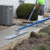 Construction worker spreads wet concrete along a wooden form for a new sidewalk beside a house, using a blue screed tool.