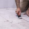 Person kneeling on a tiled floor, using a grout float to press material into grout joints near a blue bucket.