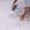 Person kneeling on light gray tile, scrubbing grout lines with a turquoise-edge grout float tool.