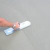 Person's arm holding a steel trowel and smoothing wet concrete surface.
