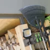 Woodpile beside a tool rack in a shed area, with a rake, shovel, and other garden tools mounted above stacked logs.